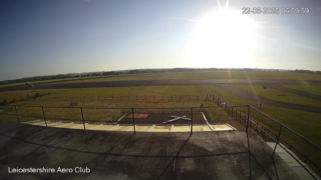 An outdoor view from a webcam at Leicestershire Aero Club showing a clear sky with the sun shining brightly. In the foreground, there is a flat rooftop with a safety railing, and beyond that, a fenced grassy area with a small white structure. Further out, there are open grassy fields and a paved runway or taxiway. The overall scene is bright with the sun positioned toward the upper right corner. The date and time are displayed in the top right corner: June 22, 2025, at 6:59:59 AM.