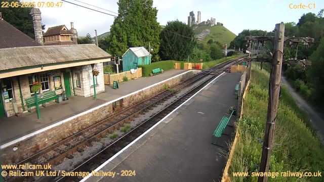 A small train station with a platform, green benches, and a building made of stone with a tiled roof. There are flower baskets hanging on lamp posts, and a small green shed in the background. Railway tracks run through the station, with a signal visible further down the tracks. Nearby, there are trees and lush greenery, with a hillside in the distance featuring ruins or an old castle on top. The image is bright, indicating early morning, with clear skies and some clouds.
