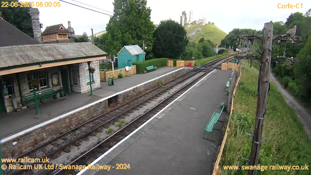 A small railway station platform with a stone building on the left, featuring a green bench and hanging flower baskets. There are train tracks running through the center, with a slightly overgrown area beside them. Several green benches are placed along the platform, and a wooden fence with flower pots borders the edge. A path runs alongside the platform to the right, with a grassy area and trees. In the background, there are green hills with a castle ruin on top, and a line of trees surrounds the station. A set of overhead electric cables runs parallel to the tracks. The timestamp shows "22-06-2025 08:00:05," and the website links are visible at the bottom.