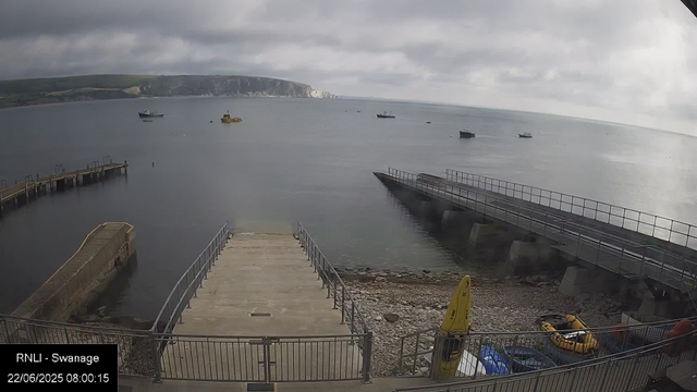 A view of a harbor with a concrete pier extending into calm water, surrounded by a metal railing. Several boats are floating on the water, and a rocky shoreline is visible in the foreground. In the background, there are white chalk cliffs and a cloudy sky. A yellow caution sign and some floating rescue equipment are near the shore. The timestamp indicates "22/06/2025 08:00:15" and the location is labeled as "RNLI - Swanage."