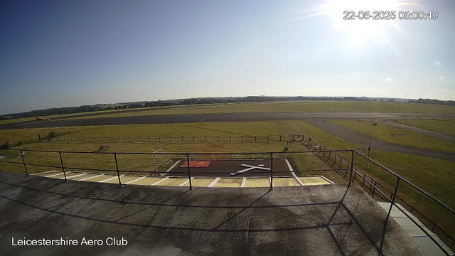 A clear view of an airfield on a sunny day, taken from a webcam positioned on a building. In the foreground, there is a fenced area with a small helipad marked with a white "H" and a red rug nearby. The grass surrounds the paved surfaces, and a flagpole with a flag is visible on the left side. The runway and taxiways extend into the distance with no aircraft visible. The sky is mostly clear with just a few small clouds, and the sun is shining brightly, causing a glare near the top right corner. The timestamp in the upper right corner shows the date and time as June 22, 2025, at 8:00 AM. The text "Leicestershire Aero Club" is visible at the bottom left corner of the image.