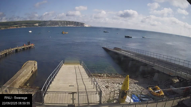 A view of a seaside harbor with a small concrete pier extending into the water. There are boats floating in the bay, including a yellow vessel. In the foreground, there is a small, fenced-off area with some equipment, including yellow life jackets and a yellow buoy. The sky is partly cloudy with white clouds scattered across a blue sky, and distant cliffs line the horizon.