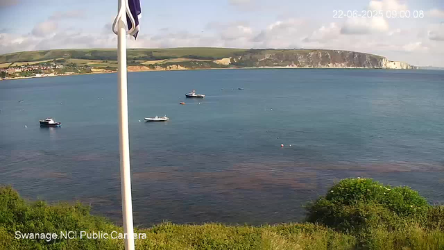 A scenic view of a calm ocean with several small boats floating on the water. In the background, there are green rolling hills and white cliffs along the coastline. The sky is partly cloudy, and a flagpole with a flag is partially visible on the left side of the image. The image is captured by the Swannage NCI Public Camera, timestamped June 22, 2025, at 09:00:08.