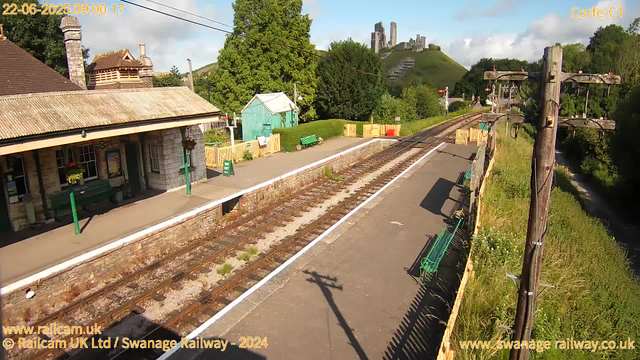 A small train station platform with a stone building on the left, featuring a chimney and a wooden roof. The platform has green benches, flower baskets, and signs. Railroad tracks run through the center, with wooden fences on either side. Overhead, utility poles with wires stretch across the scene. In the background, greenery and trees surround a hilltop with historic stone ruins. The sky is partly cloudy, and bright sunlight casts shadows on the platform. The scene is calm and appears to be early morning.