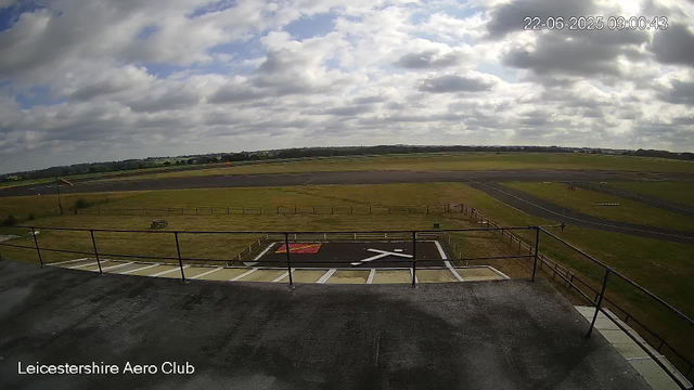An outdoor view from a webcam at Leicesterhire Aero Club showing a cloudy sky over a runway and grassy fields. There is a helipad with a red and white symbol in the foreground, and a small field with a fence and a flag will waving on the left side. The landscape extends into the distance with a few structures visible on the horizon.
