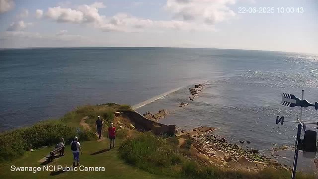 A coastal scene with a grassy area in the foreground where four people are walking or sitting, some wearing hats and backpacks. A stone path extends into the ocean, surrounded by rocks. The ocean stretches out with gentle waves, and the sky is partly cloudy with sunlight reflecting off the water. The image has a timestamp in the upper right corner and text in the lower left corner that reads "Swanage NCI Public Camera."