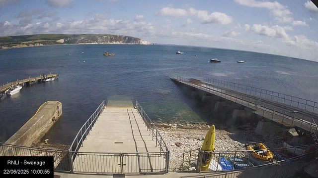 A view of a harbor with a concrete pier extending into the water, surrounded by metal railings. Several small boats are moored along the piers, and a few boats are sailing in the distance. In the foreground, there are yellow lifesaving equipment and a yellow buoy on a dock area with rocks and gravel. The background features a cliffside with white chalk cliffs and a partly cloudy sky. The timestamp indicates the photo was taken on June 22, 2025, at 10:00 AM.
