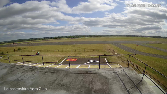 A view from a webcam at Leicestershire Aero Club shows a runway and grassy airfield under a partly cloudy sky. In the foreground, there is a rooftop with a railing, a helicopter parking area painted with red and white markings, and a small vehicle parked on the grass. The background features a large open field with a long runway, and a line of trees and distant fields on the horizon. The timestamp in the upper right corner reads "22-06-2025 10:00:59."