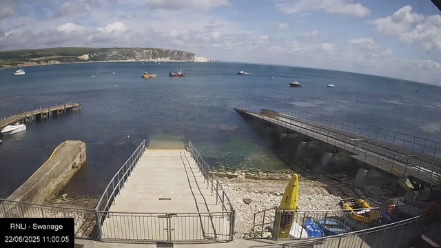 A view of a coastal dock with boats anchored in the water. There are two piers extending into the sea, with a small boat on the left dock. The sky is partly cloudy, and in the background, there are white chalk cliffs and green hillside. A yellow buoy and some rescue equipment are visible near the dock. The timestamp reads "22/06/2025 11:00:05."