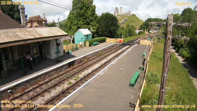 A small rural train station platform with a stone building on the left, where two people are standing and talking. The platform is lined with green benches and decorative planters. Railway tracks run through the center, with a safety fence at the far end. Behind the station, there are lush green trees and a hill with what appears to be a castle ruin at the top. To the right, there are tall wooden utility poles with electrical wires, and a narrow footpath runs alongside the station. The sky is partly cloudy, and the scene is well-lit, indicating a bright day. The image is marked with the date and time: June 22, 2025, at 10:59 AM.