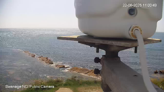 A webcam captures a view of the ocean with sunlight reflecting off the water. In the foreground, part of a device with a white container and a hose connected to it is visible, mounted on a metal stand. The scene is bright and clear, with a rocky shoreline extending into the water. The timestamp in the upper right corner shows the date and time as June 22, 2025, 11:00:49. Text at the bottom indicates the camera is a Swannage NCI Public Camera.