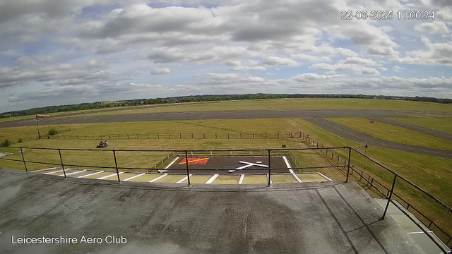 A wide view of an airfield taken from a webcam at Leicestershire Aero Club. In the foreground, there is a small parking area with white lines and a red marked space. A fence runs across the image, with a small flagpole to the left. Two people are sitting on a bench on the grass. The runway stretches into the distance under a partly cloudy sky, with green fields and trees visible on the horizon. The date and time in the upper right corner indicate June 22, 2025, at 11:00:34.