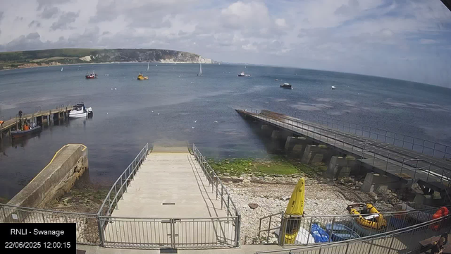 A scenic view of a harbor with several boats floating on calm waters, a dock extending into the sea, and a rocky shoreline. In the background, there are green hills with white cliffs under a partly cloudy sky. The foreground shows a small concrete pier with railings, some yellow safety equipment, and a few paddle boats on the rocky beach area.