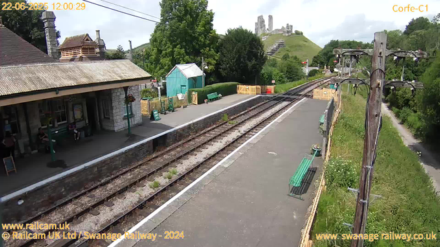 A railway station platform with a small shelter on the left, where a few people are sitting on benches. There are green benches, flower pots, and some signs. The tracks run through the middle, with an electric pole on the right side. In the background, there is green foliage, a small building, and a hill with ruins or an old castle on top. The sky is partly cloudy with patches of sunlight. The timestamp reads "22-06-2025 12:00:29".
