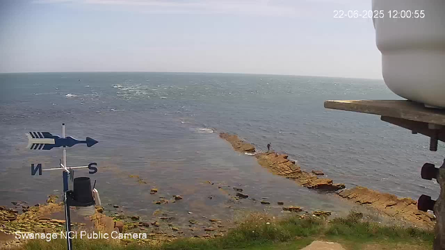 A view of a seaside landscape with a rocky shoreline extending into the ocean. The water is calm, with small ripples, and the sky is clear. There is a person walking along the rocks in the distance. In the foreground, a weather vane with directional markers (N, S) is mounted on a pole, and part of a large white container is visible on the right side. The top right corner shows a timestamp reading "22-06-2025 12:00:55".