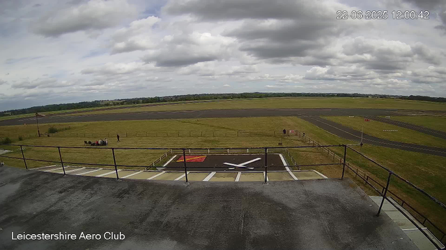 The image shows an outdoor view from a webcam at Leicesterhire Aero Club. In the foreground, there is a small helicopter landing pad with a red and white marked area and a single helicopter parked on it. A black railing surrounds the area. Beyond the landing pad, there is a grassy field with a few people sitting on benches and walking. In the distance, a runway extends across the image, with a cloudy sky overhead. The timestamp in the top right corner indicates the date and time as June 22, 2025, at 12:00:42.