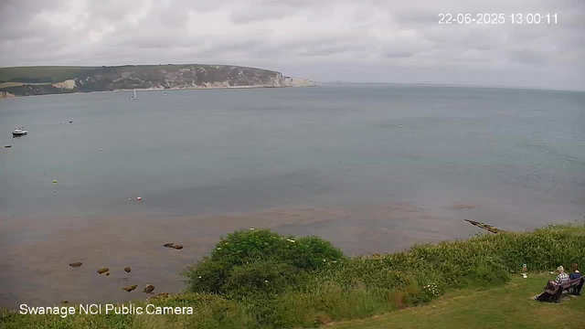 A view of a calm bay with a few boats floating on the water. In the background, there are white chalk cliffs with green fields on top. The sky is overcast with gray clouds. In the foreground, there is a grassy area with some rocks near the water and a bush. On the right side, two people are sitting on a bench facing the sea. The timestamp reads "22-06-2025 13:00:11" and the image is labeled "Swanage NCI Public Camera."