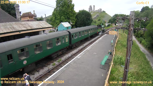 A vintage steam train with dark green carriages is arriving at a rural station platform. The steam locomotive is black with steam puffing out, pulling the carriages along the track. The platform is bordered by a wooden fence with potted plants and teal-colored benches. A utility pole with electrical wires runs along the right side, and there is a narrow path with grass and trees beyond the fence. In the background, a hill with ruins of a castle or fortress can be seen, alongside some buildings with stone roofs. The sky is overcast, and the timestamp indicates the photo was taken on June 22, 2025, at 1:00 PM.