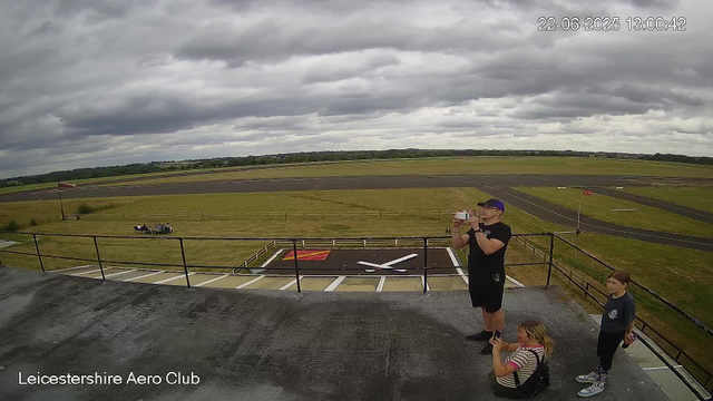 A group of three people, one adult man and two young girls, are standing on a platform at Leicesterhire Aero Club. The man is taking a photo or video with a smartphone. One girl, with blonde hair and wearing a striped shirt, is crouching and holding a phone, possibly recording. The other girl, standing and holding a drink, has dark hair and is dressed in a dark shirt and patterned leggings. The scene is set outdoors on a cloudy day, with a grassy field and a runway visible in the background. There is a small wind sock on a pole and a couple of small aircraft decals on the ground near the platform. The timestamp shows the date as June 22, 2025, and the time as 13:00:42. The location is labeled as Leicesterhire Aero Club.