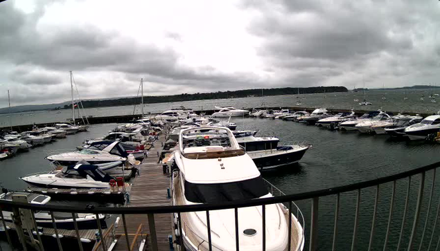 A marina filled with numerous boats and yachts docked along piers, with a cloudy sky overhead and the open water extending into the distance.