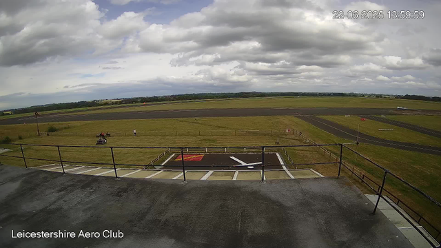 This image shows a view from a webcam at Leicestershire Aero Club. In the foreground, there is a small parking area with white lines and a designated helicopter landing pad marked with a red square and a white helicopter symbol. A metal railing surrounds the area. Beyond, a grassy field contains a few small vehicles and several people. Further in the background, there is a runway, some distant buildings, and a landscape of fields and hills under a cloudy sky. The timestamp in the upper right corner indicates the date and time as June 22, 2025, at 13:59:59.