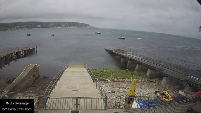 A view of a harbor with boats floating on calm water, a concrete pier extending into the sea, and a shoreline with rocks and grass in the foreground. The sky is overcast with gray clouds. In the background, there are cliffs and hills. A yellow life raft and other equipment are visible near the fence at the bottom right corner. The timestamp indicates the date and time as June 22, 2025, at 14:00.