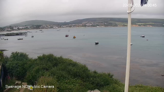 A view of a harbor with several boats anchored on calm water. In the background, there are hills with some buildings and greenery. The sky is overcast, and the scene is captured from a webcam, with the timestamp showing August 22, 2023, at 14:00:45. In the foreground, there is some lush green foliage and a partial view of a white pole. The bottom left corner has the text "Swanage NCC Public Camera."