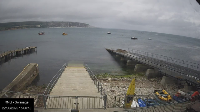 A view of a cloudy day at a coastal area with a concrete pier and a ramp leading into the water. Several boats are anchored in the bay, and a distant headland with cliffs is visible across the water. There are some small construction or safety equipment items, including a yellow caution sign and a yellow inflatable boat, near the shore. The timestamp indicates the photo was taken on June 22, 2025, at 3:00 PM.
