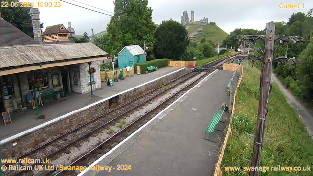 A small train station platform with two people sitting on a bench beneath a stone building. There are green benches, flower pots, and a small teal-colored shed near the station. Railroad tracks run through the middle of the image. On the right, there are wooden utility poles with wires overhead. In the background, a lush green hillside rises with an old castle ruin on top. The sky is overcast.