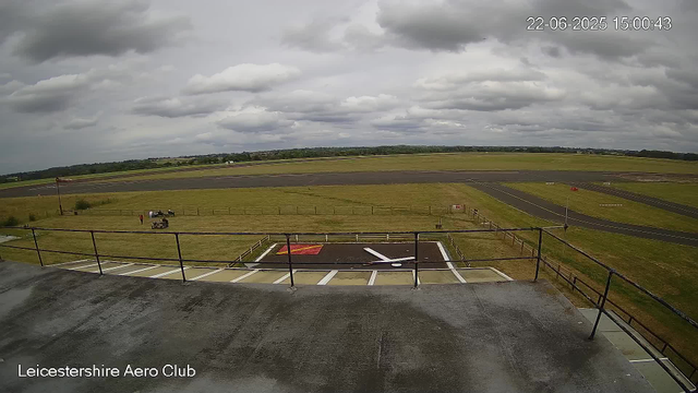 A view from a webcam at Leicestershire Aero Club showing an overcast sky with dark clouds. In the foreground, there is a platform with a railing, a marked helipad with a white helicopter symbol, and airplanes parked nearby. Beyond, there is a grassy area with a few people and vehicles, and a runway extending into the distance. The landscape is mostly flat with some trees visible on the horizon.