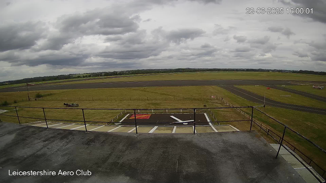 A view from a webcam at Leicesterhire Aero Club shows an overcast sky with dark, heavy clouds. In the foreground, there is a paved platform with safety railings and a designated helicopter landing pad marked with a red and yellow symbol. A small helicopter is parked on the grass nearby, and there are multiple runways and taxiways stretching into the distance. The landscape is flat with fields and some distant trees under a cloudy sky. The timestamp indicates the photo was taken on June 22, 2025, at 16:00.