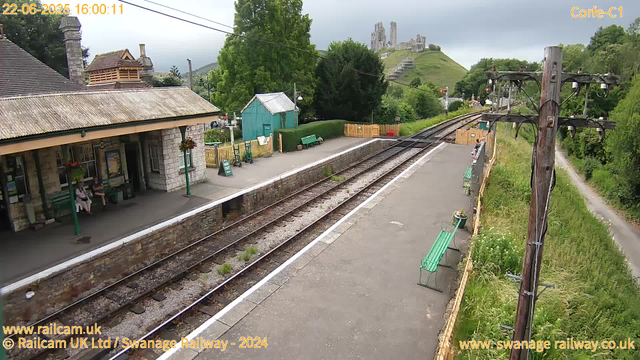 A small railway station platform with a stone building on the left, where two people are sitting on a green bench. There are a few flower baskets hanging from the building. The platform has a few green benches, a small green shed, and some plants near the fence. Railway tracks run through the center of the image. On the right side, there is a wooden utility pole with electrical wires. In the background, a green hillside with a castle ruin on top is visible, along with trees, a walking path, and a cloudy sky.