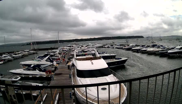 A marina filled with numerous boats and yachts docked along wooden piers, with overcast skies and dark clouds overhead. The water is calm, and there are several sailboats and motorboats lined up, with some people visible on the docks. In the background, a distant shoreline and hills are visible under the cloudy sky.