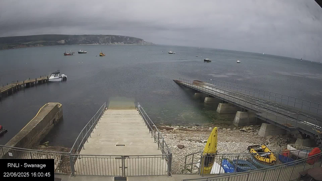 A view of a harbor on a cloudy day with several boats anchored in the water. There is a concrete pier extending into the water from the bottom left, with a small boat moored to it. To the right, a metal ramp leads down to the rocky shoreline. In the foreground, there are yellow safety barriers, a small inflatable boat, and some blue crates. The background features a distant coastline with cliffs under a dark, overcast sky.