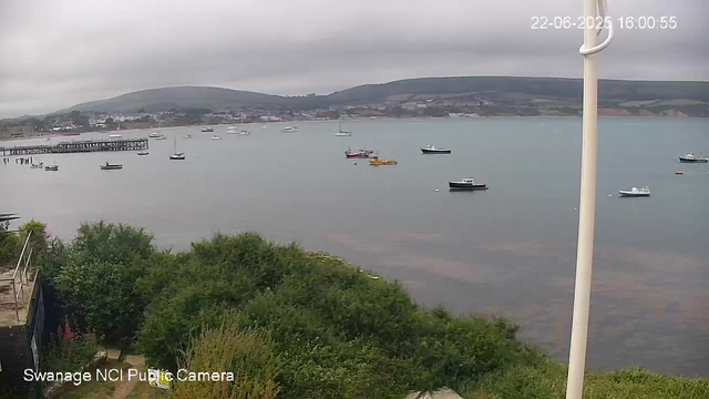 A view of a harbor with several boats floating on calm water. In the background, there are rolling green hills under a cloudy sky. In the foreground, there is a small area with green bushes and a pathway, partially visible, along with a white pole on the right side of the image. The timestamp in the upper right corner reads "22-06-2025 16:00:35" and the text at the bottom left states "Swanage NCI Public Camera."