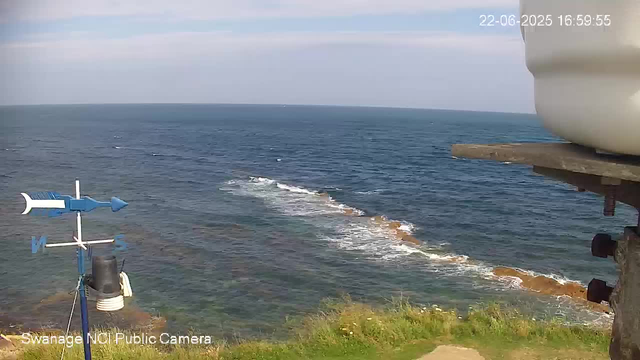 A view of the ocean with gentle waves and a distant horizon under a partly cloudy sky. In the foreground, there is a weather vane and a small satellite dish mounted on a structure near the shoreline. The scene appears to be captured from a webcam on a clear day.
