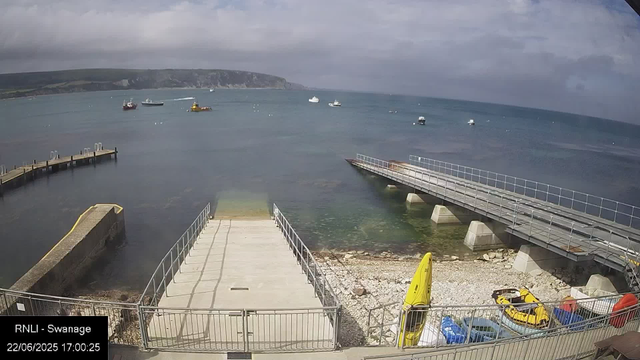 A view of a waterfront with a concrete and metal pier extending into the sea. Several small boats are anchored in the water, and a yellow rescue boat and some equipment are on the shore. The sky is cloudy, and a distant coastline with cliffs is visible across the water. The timestamp shows "22/06/2025 17:00:25" and the location label reads "RNLI - Swanage."