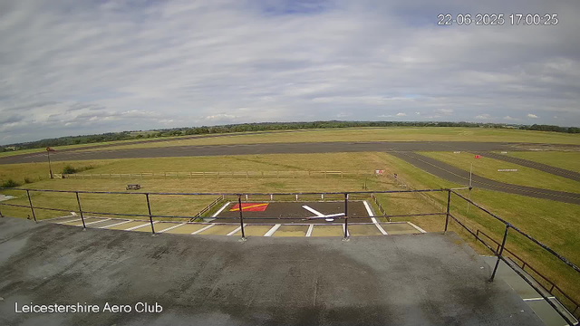 A wide view of an airfield with taxiways and runways under a partly cloudy sky. There is a fenced area with parking spaces and a helipad marked with a red and yellow symbol. In the background, open grassy fields stretch out towards a distant tree line. The timestamp on the image reads "22-06-2025 17:00:25" and the bottom left corner indicates it is from "Leicestershire Aero Club."