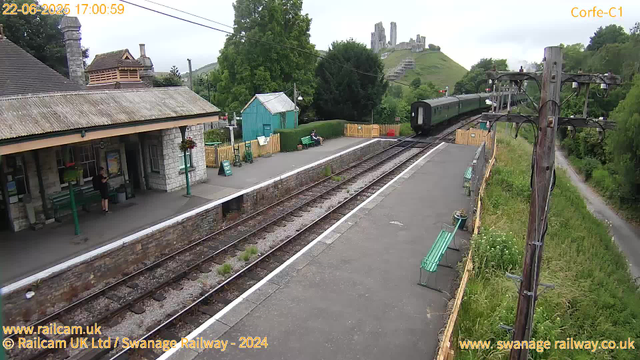 A small train station platform with a single green train approaching from the distance. On the left side, there is a stone building with a sign that says "Swansea" and a small covered waiting area with green benches. A person is standing under the shelter, and a couple of benches are positioned along the platform. Near the platform, there are a few small shrubs and flower pots hanging from posts. On the right side, a tall wooden utility pole with wires runs along the green pathway parallel to the railway tracks. Lush green trees and a grassy hill with a castle ruin on top are visible in the background. The sky is cloudy, and the overall scene appears calm and quiet. The timestamp in the top left corner indicates the date is June 22, 2025, at 17:00.