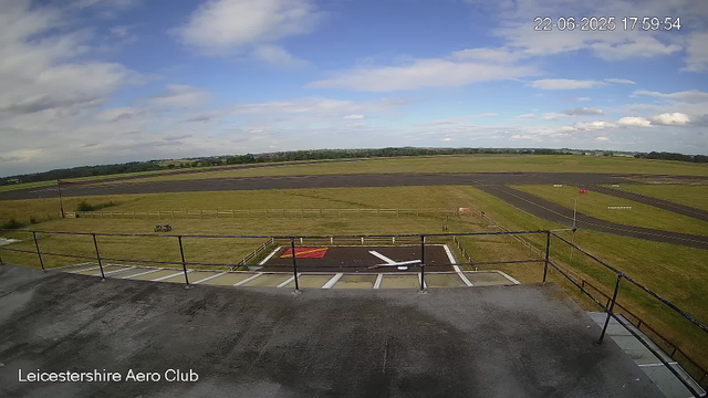 A wide view of an airport runway and surrounding grassy fields under a partly cloudy sky. In the foreground, there is a helipad marked with a white "H" and red borders, enclosed by a black railing. The image shows the Leicestershire Aero Club, with a timestamp in the top right corner indicating it was taken on June 22, 2025, at 17:59:54.