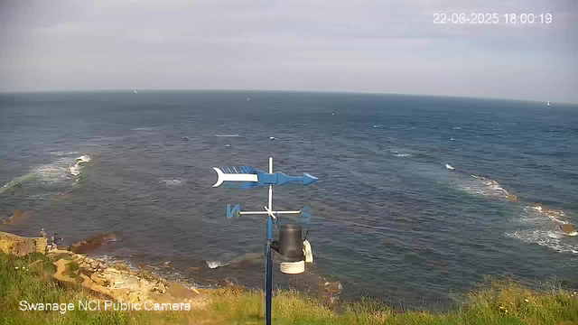 A scene of the ocean viewed from a webcam, with waves and small boats in the distance. In the foreground, there is a weather vane mounted on a pole pointing to the north. The sky is partly cloudy, and the timestamp in the upper right corner reads "22-06-2025 18:00:19."