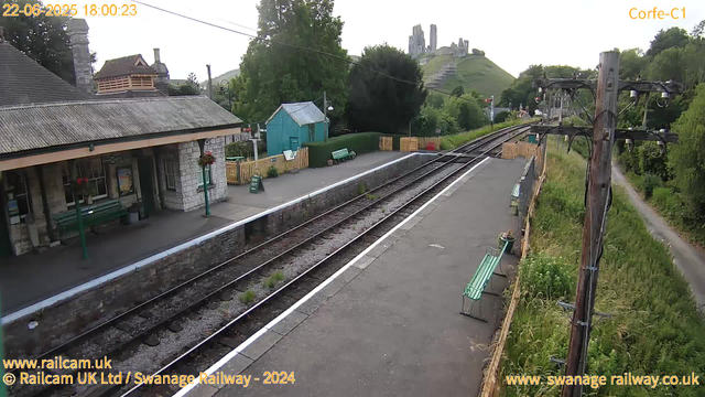 A small train station platform with a stone building on the left, green benches, and a small blue shed behind a yellow fence. There are four wooden platform barriers and a sign that says "Way Out." To the right, there is a utility pole with wires and two benches along the fence. Railway tracks run through the center, curving off into the distance. In the background, a hill with what appears to be an ancient castle ruins on top is visible, surrounded by trees and greenery. The timestamp indicates the image was captured on June 22, 2025, at 6:00 PM.