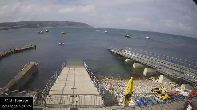 A view of a harbor with several boats floating on the water. There is a concrete pier extending into the water in the foreground, with a smaller dock to the left. To the right, a metal ramp leads down to the shore, which is covered with rocks and pebbles. Two yellow kayaks or paddleboards are on the ground near the ramp, and a few cars are parked behind a fence at the bottom of the image. In the background, there are rolling hills under a partly cloudy sky. The timestamp indicates the date and time as June 22, 2025, 6:00 PM.