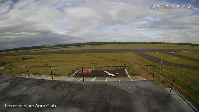 An outdoor view from a webcam at Leicesterhire Aero Club shows a paved area with a railing at the edge, a small marked landing pad, grassy fields, a runway in the distance, and a partly cloudy sky overhead. The timestamp in the top right corner reads "22-06-2025 18:59:58".
