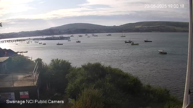 A view of a bay with several boats anchored on the water. In the background, there are hills and a cloudy sky. The timestamp in the top right corner reads "22-06-2025 19:00:18". The bottom left corner indicates it is from the "Swanage NCI Public Camera."
