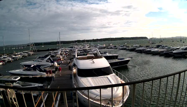A view from a webcam showing a marina filled with numerous boats and yachts docked along wooden piers. The sky is partly cloudy, and the sea extends to the horizon with more boats anchored in the distance. A person in red clothing is walking along the dock near the center of the image. A metal railing is visible in the foreground.