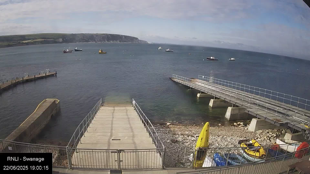 A view of a harbor with a concrete pier extending into the water, surrounded by metal railings. There is a yellow kayak and some equipment stored on the rocky shore below. Several boats are anchored in the water, and green rolling hills with cliffs are visible in the background. The sky is partly cloudy with patches of blue. The timestamp indicates the date as June 22, 2025, at 19:00.