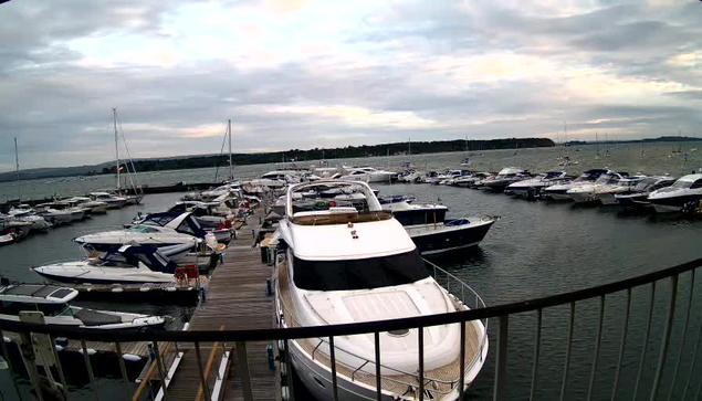 A marina filled with numerous boats and yachts docked along wooden piers under a cloudy sky. A railing is visible in the foreground, and the water appears calm with some gentle waves. The background shows a distant shoreline with trees and a horizon line.