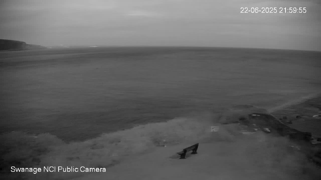 A black-and-white image from a webcam shows a beach scene with a bench in the foreground near the shoreline. The ocean stretches into the distance with waves gently lapping the shore. In the background, there is a coastline with low cliffs or hills on the left side. The sky appears mostly cloudy. The timestamp in the top right corner reads "22-06-2025 21:59:55," and the bottom left corner indicates this is a "Swanage NCI Public Camera."