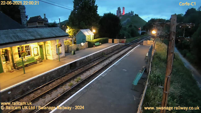 A small train station platform at dusk or early evening with a historic building illuminated by yellow lighting on the left. There are benches, potted plants, and hanging flower baskets along the platform. The railway tracks run through the center of the image, with some vegetation growing between the sleepers. To the right, there are utility poles with wires running alongside the tracks and a grassy embankment. In the background, a hill with ruins or a castle remains overlooking the station, and there is a partly cloudy sky. The scene is peaceful, with no people visible.